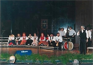 Grupo de músicos e cantores tradicionais em palco, sentados e em pé, com instrumentos e trajes típicos, durante uma atuação cultural.