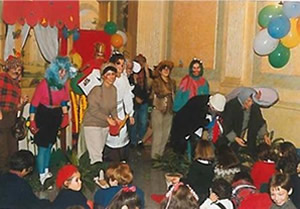 Grupo de adultos e crianças em palco, vestidos com fatos coloridos e fantasias, durante uma festa infantil com balões e decoração alegre.