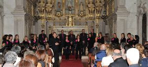 Grupo coral em palco dentro de uma igreja, vestidos maioritariamente de preto com detalhes em rosa, perante uma plateia sentada.