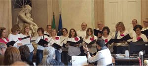 Grupo coral a cantar em sala institucional, segurando partituras, com público sentado em primeiro plano.